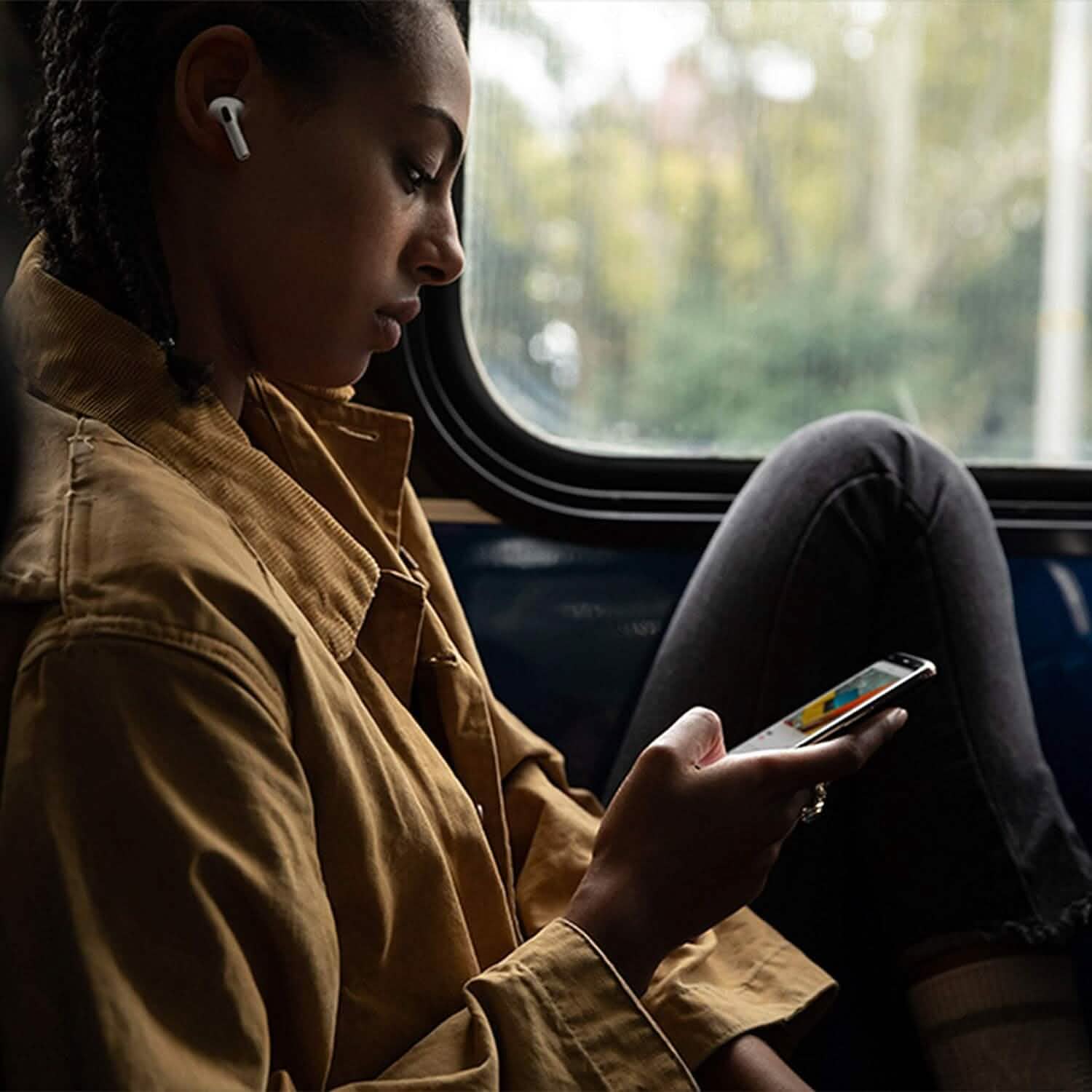 Wearing a brown jacket and using Apple AirPods Pro (1st Gen) Refurbished — Right, Left, or Charging Case, a person sits by a bus window with one knee bent, looking at their smartphone as daylight streams in.