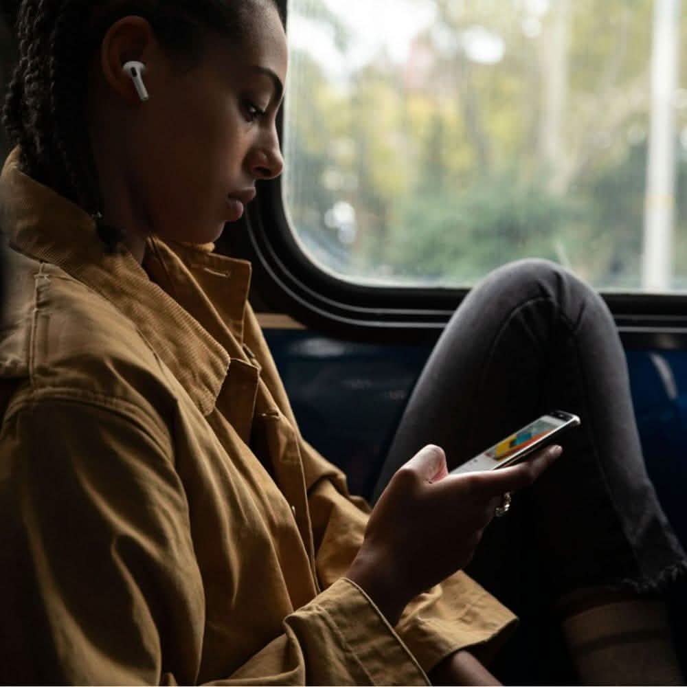 A person in a brown jacket sits by a window on a bus or train, wearing Apple AirPods Pro with Wireless Charging Case and looking at their phone. Trees and blurred scenery are visible outside.