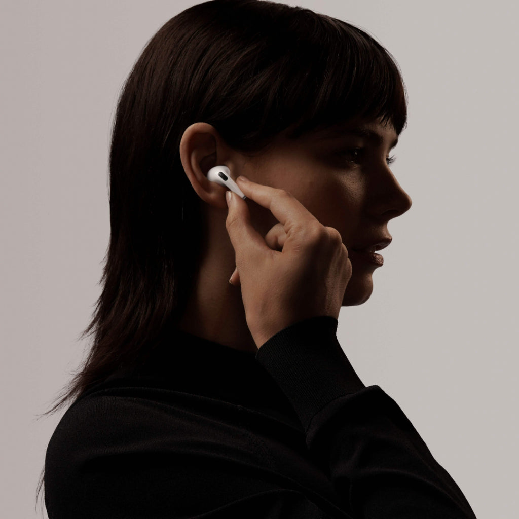 A person with short dark hair wears a black top and inserts an Apple AirPods Pro with Wireless Charging Case earbud, featuring Active Noise Cancellation, into their ear, holding it with one hand against a neutral background.